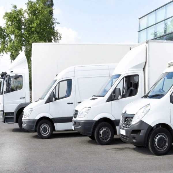 Five white commercial vehicles parked in a diagonal row outside a modern gray industrial building. The fleet ranges from small delivery cargo vans to larger box trucks and a semi-truck cab on the far left.